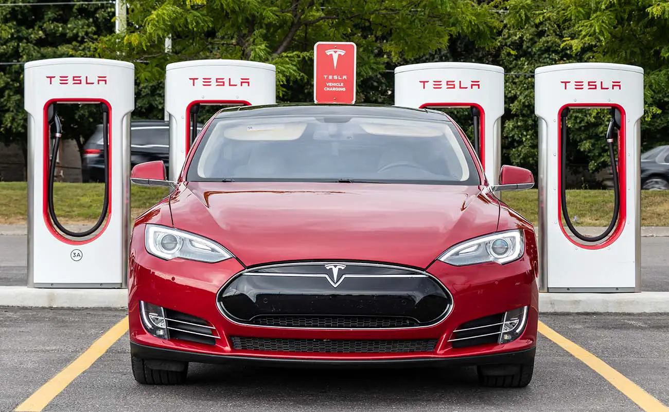 red tesla model s parked in front of tesla supercharger station with busy road in background